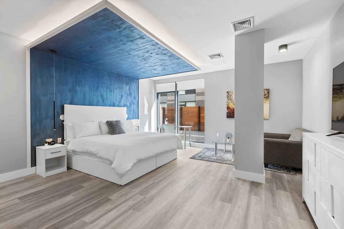 Une chambre moderne avec un grand lit, une table de chevet, une commode et une vue sur le salon grâce à une porte vitrée.