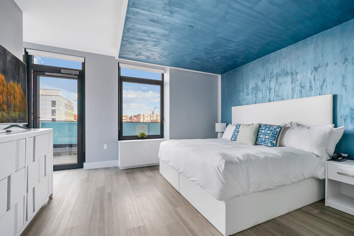 Une chambre moderne avec un grand lit, une table de chevet, une fenêtre donnant sur la ville et une porte menant à un balcon.