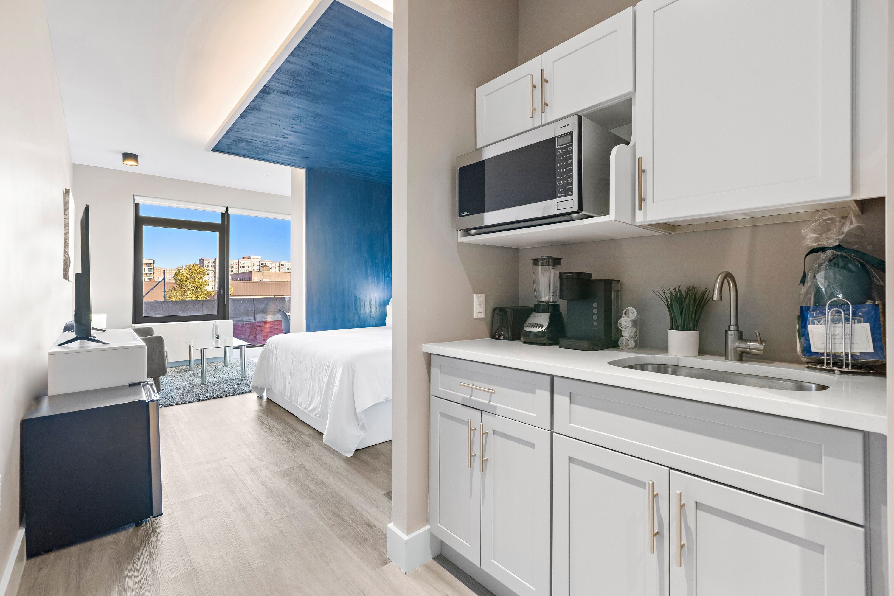 A modern hotel room with a kitchenette, a bed with white bedding, a desk with a computer, and a view of the city through large windows.