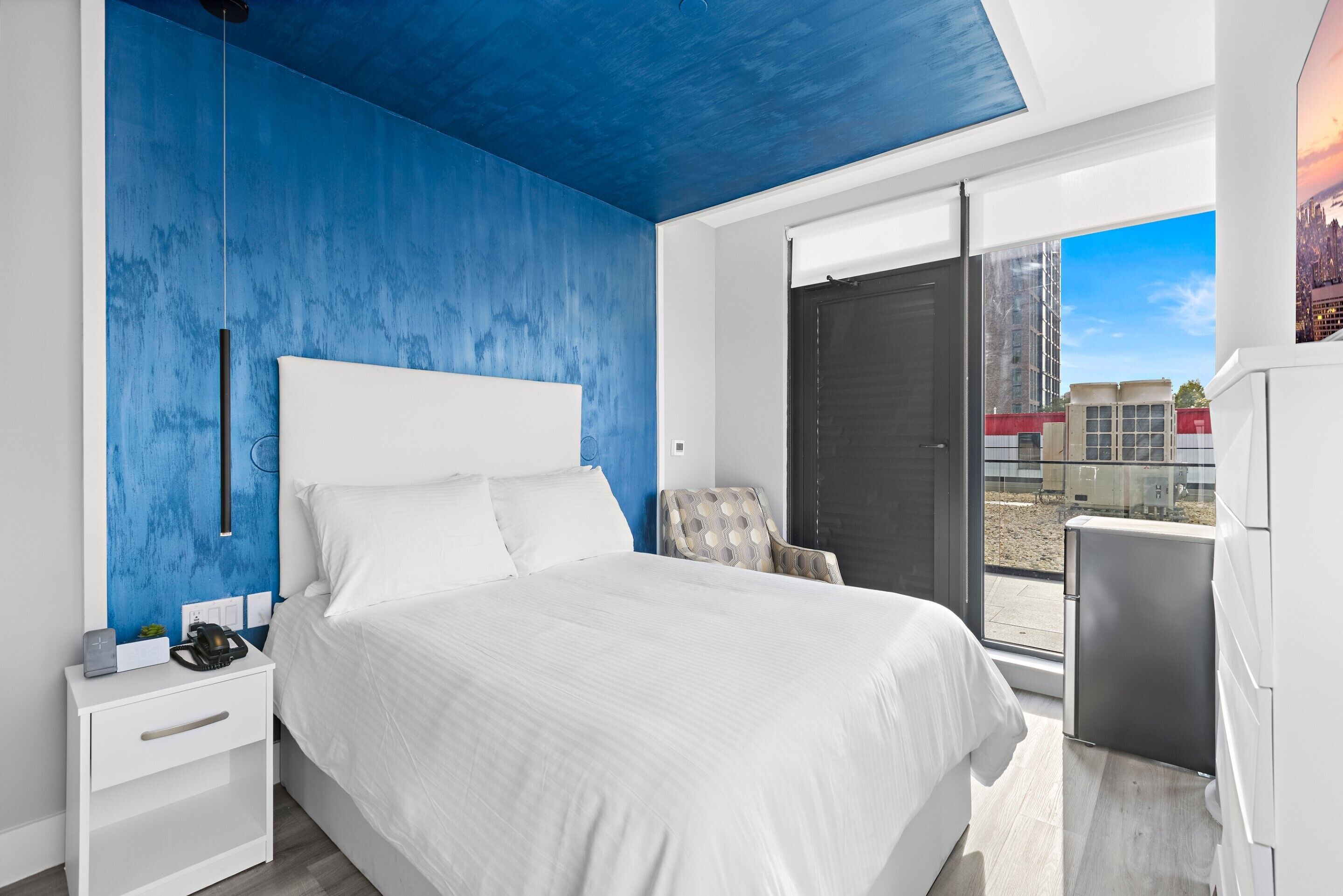 Un dormitorio moderno con una cama grande, una mesita de noche, una silla y vistas de la ciudad a través de una puerta corredera.