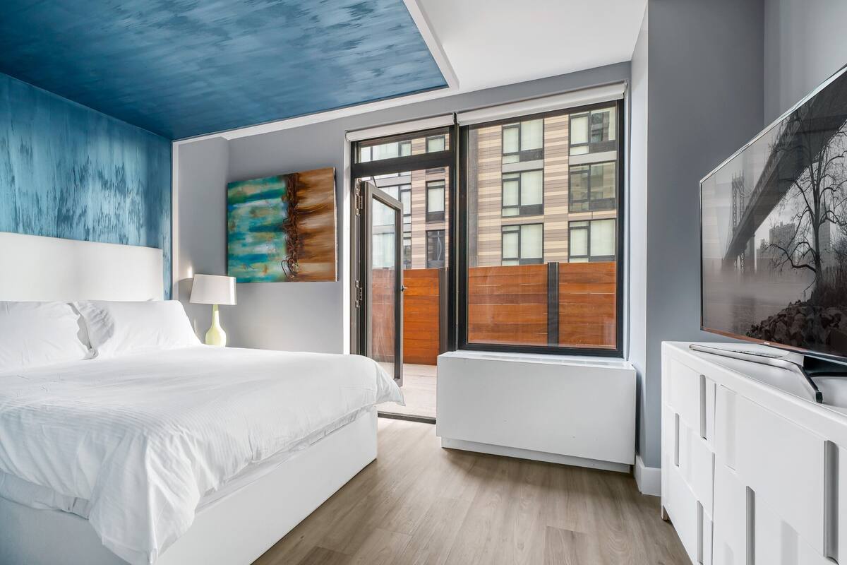 Une chambre moderne avec un lit, une fenêtre donnant sur des immeubles, un tableau au mur et un téléviseur posé sur un meuble.