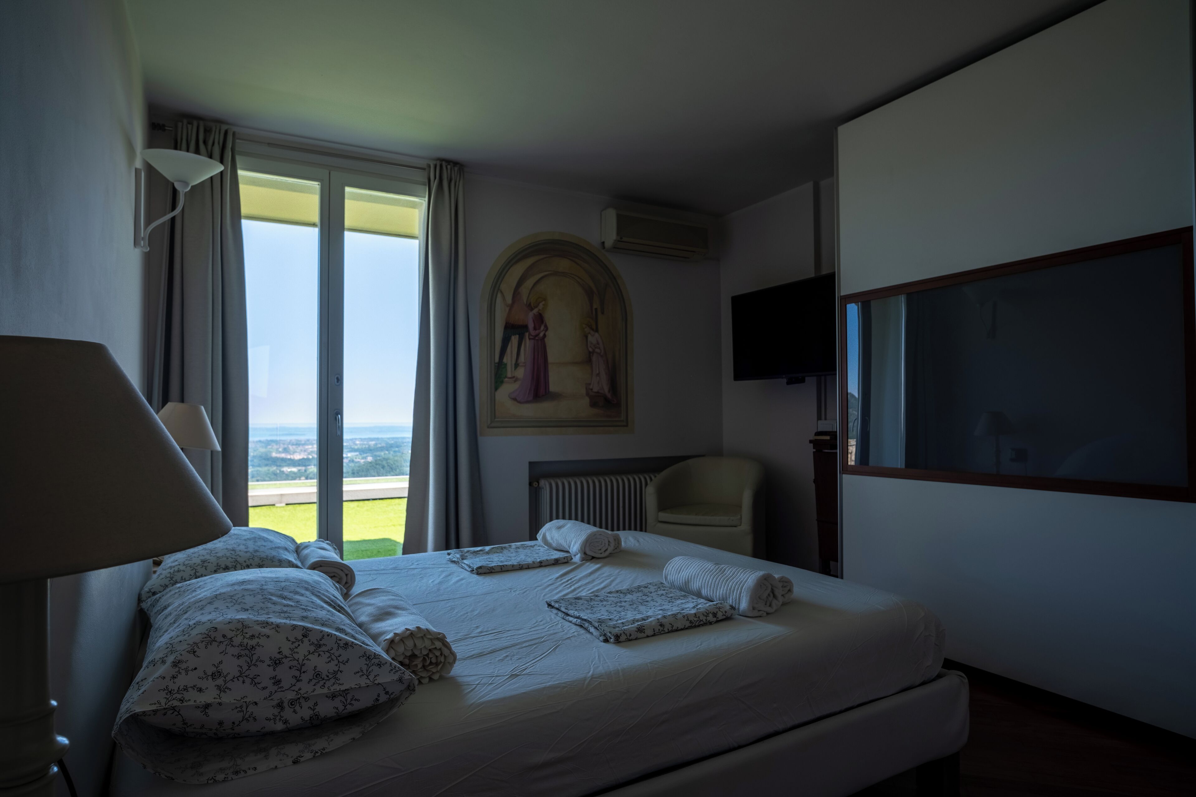 A hotel room with a large bed, a chair, a TV, and a painting on the wall.