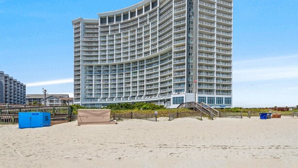 Un hotel grande de varios pisos con una fachada curva, situado en una playa arenosa con una valla de madera y una escalera que lleva a la entrada.
