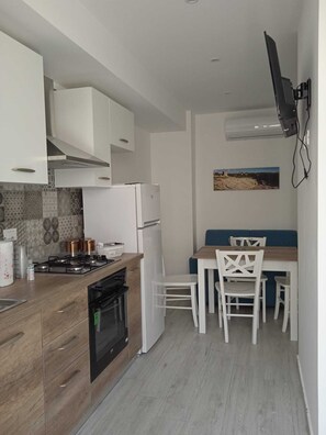 A compact kitchen with white cabinets, a gas stove, and a built-in oven. There is a dining area with a table and chairs, and a wall-mounted TV. - Ambra apartment (Avola)