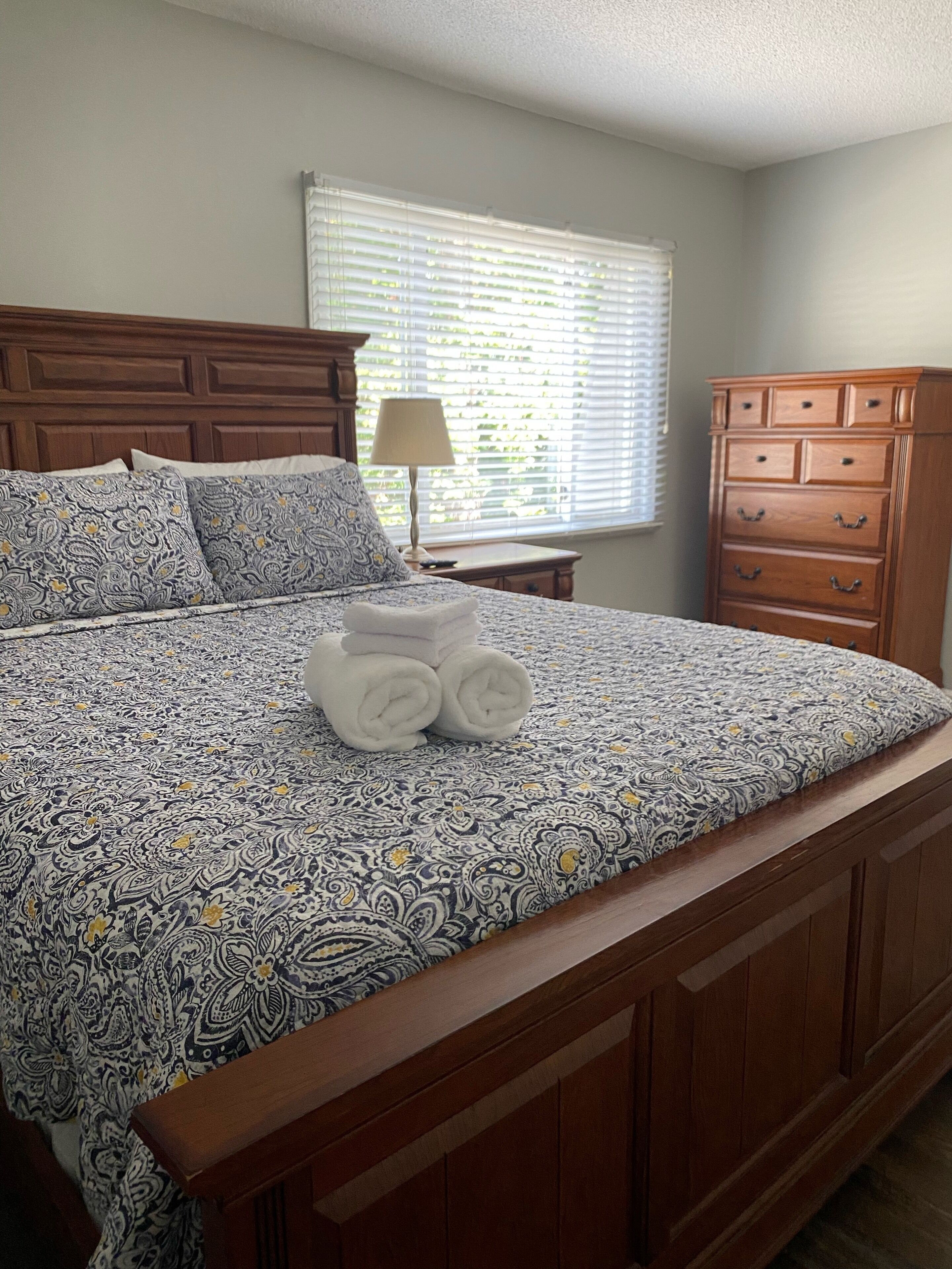 Una cama bien hecha con ropa de cama estampada, una lámpara en una mesita de noche y un aparador de madera en un dormitorio.