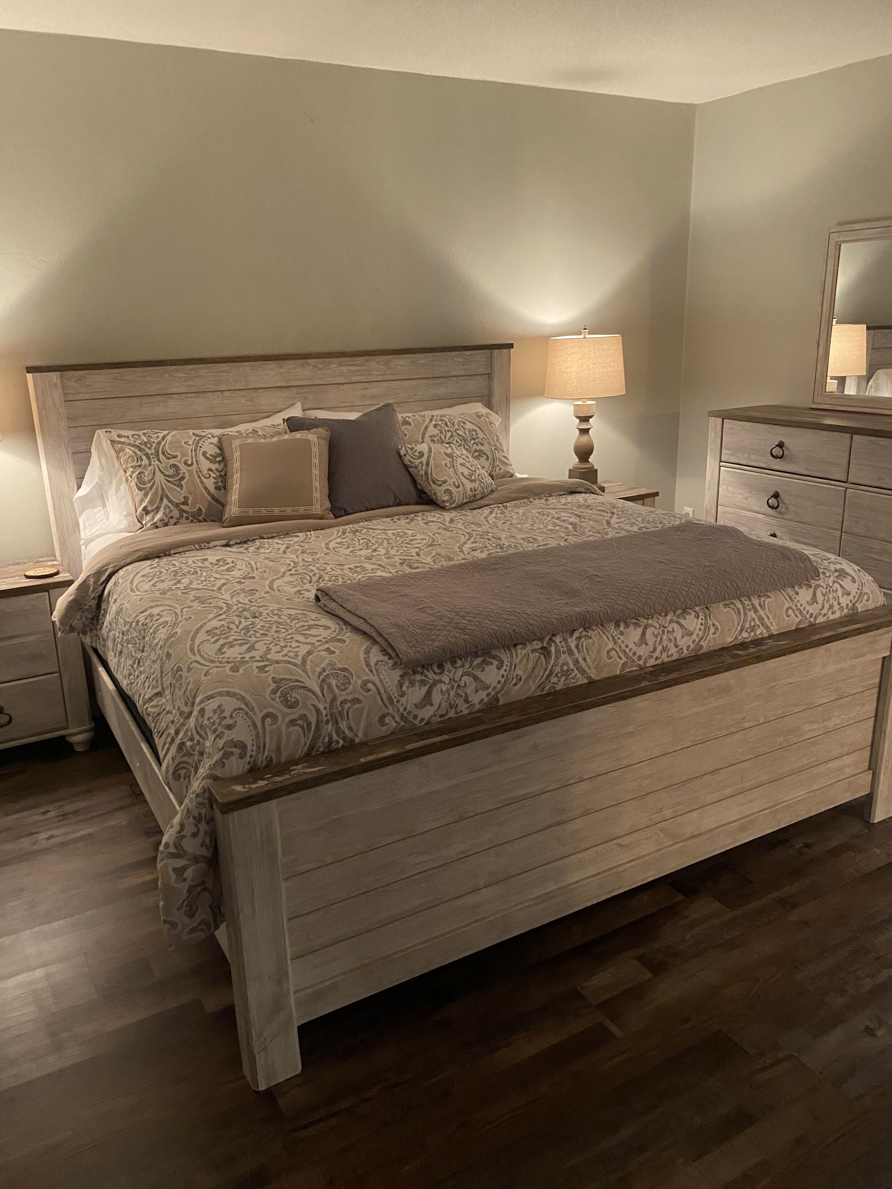 Een slaapkamer met een houten bedframe, een bed met een gedessineerd sprei, twee bedlampen en een nachtkastje met een lamp.