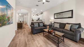 Un salon moderne avec un canapé gris, une table basse et un ventilateur de plafond. On aperçoit une cuisine en arrière-plan, avec des meubles blancs et un grand miroir au mur.