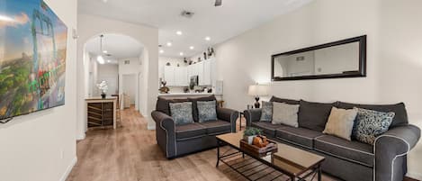 A modern living room with a grey sofa, a coffee table, and a ceiling fan. There is a kitchen in the background with white cabinets and a large mirror on the wall.
