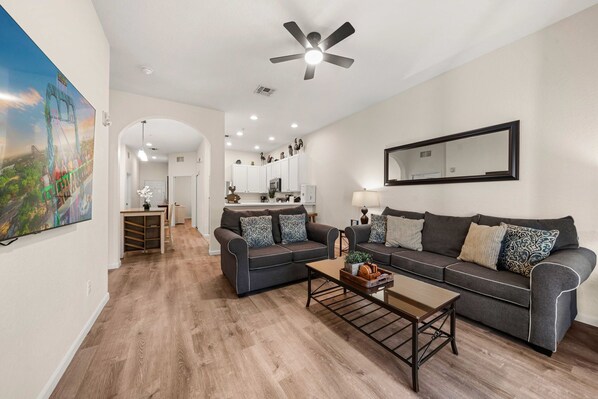 A modern living room with a grey sofa, a coffee table, and a ceiling fan. There is a kitchen in the background with white cabinets and a large mirror on the wall.