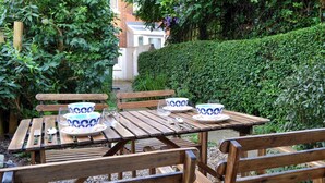 Une table en bois, dressée avec des assiettes, des bols et des couverts, entourée de bancs dans un jardin à la végétation luxuriante, avec une haie en arrière-plan.