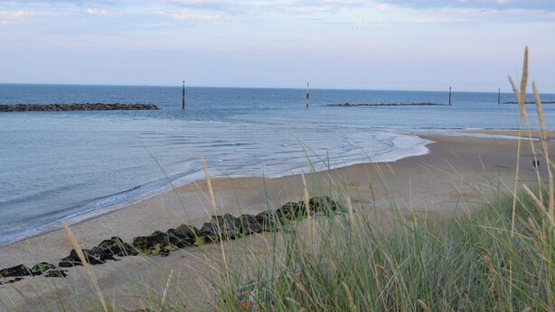 Une plage aux eaux calmes, une digue rocheuse et des dunes herbeuses.