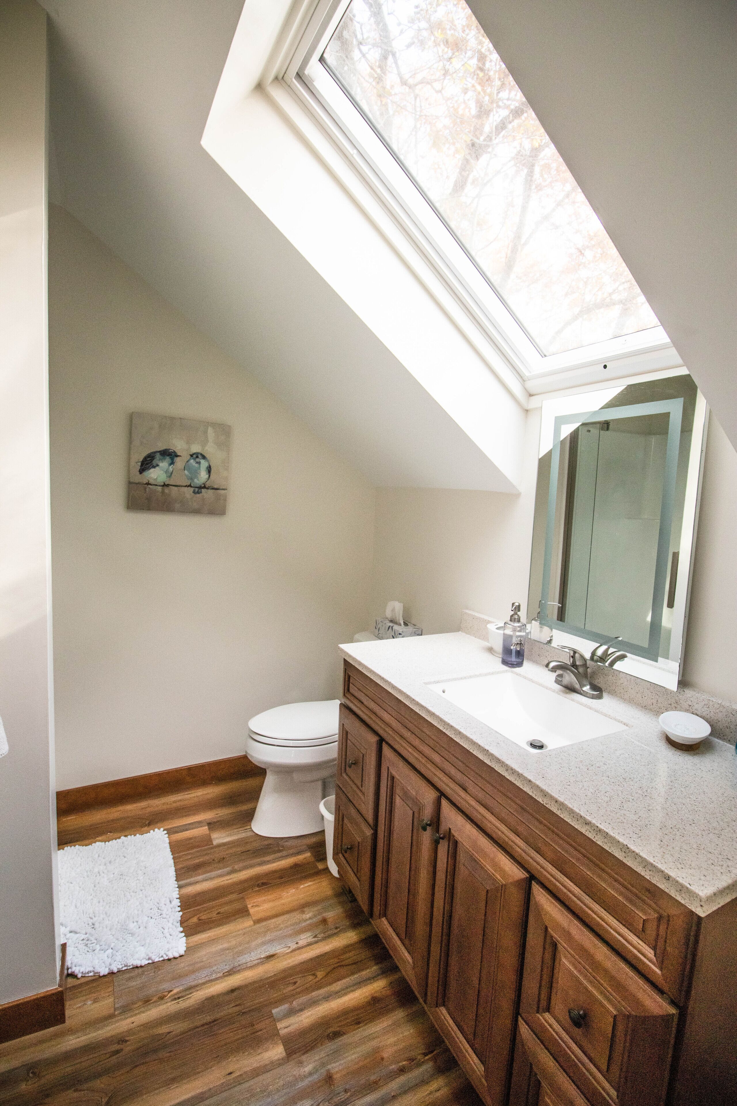 Un baño con tragaluz, piso de madera, inodoro blanco y tocador de madera con espejo.
