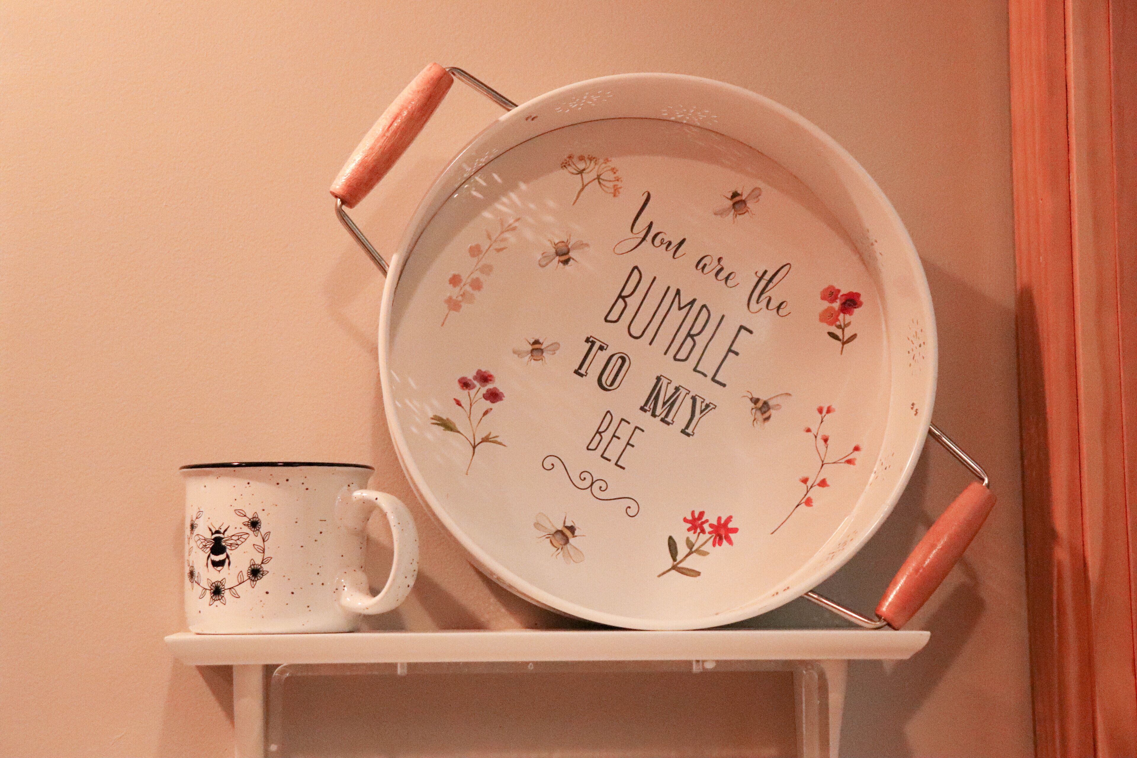 A kitchen shelf with a white mug and a white pan with floral designs and text.