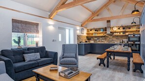 A modern living room with a sofa, armchair, and coffee table, a kitchen with wooden beams and brick walls, and a dining area with a wooden table and benches. - 3 bedroom accommodation in Llanboidy (Llanboidy)