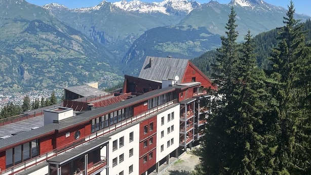 Hotel com fachada vermelha e branca, várias varandas e montanhas ao fundo.