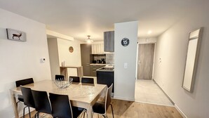 Cozinha moderna com armários de madeira e uma ilha central. Há uma área de refeições com mesa e cadeiras, e um corredor que leva a uma porta com o número 15.