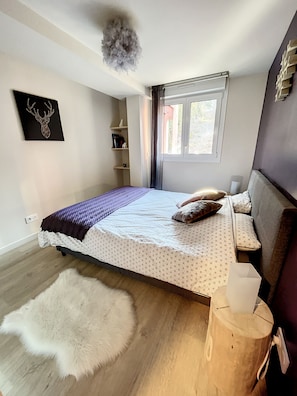 A bedroom with a bed, a white furry rug, a wooden nightstand, a window with curtains, and a deer head artwork on the wall.