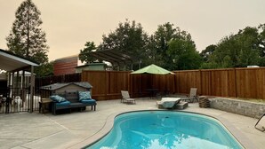 A backyard with a swimming pool, patio furniture, a covered seating area, and a wooden fence. - Spacious Custom Retreat with a Pool! (Central Point)