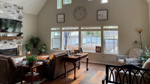A spacious living room with a stone fireplace, a large TV, and multiple windows offering views of the outdoors. - Spacious Custom Retreat with a Pool! (Central Point)