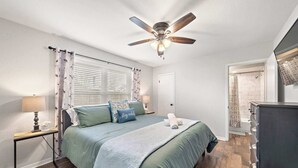 Une chambre à coucher avec un lit, un ventilateur de plafond, une table de chevet avec une lampe, une commode et une porte de salle de bain.