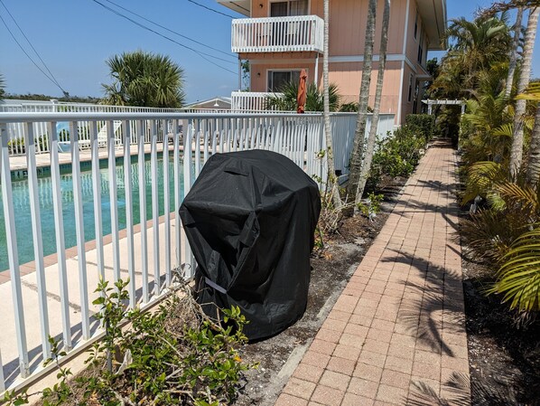 A covered grill near a poolside with a white fence, palm trees, and a building in the background.