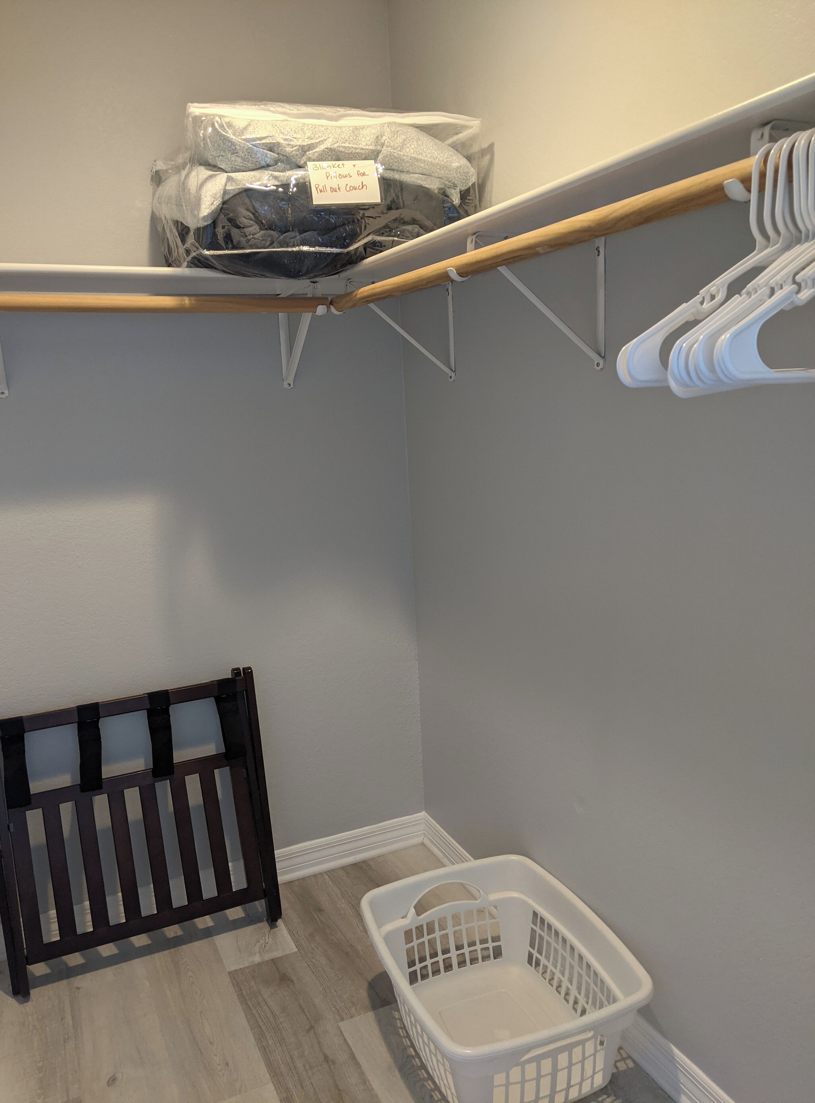 A laundry room with a wooden floor, a white laundry basket, a black radiator, and a storage area with a wooden rod and white hangers.