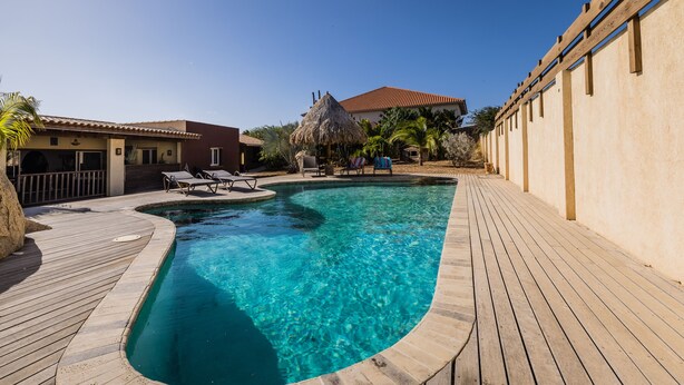 A clear swimming pool with a wooden deck surrounding it, a thatched-roof structure, and lounge chairs.