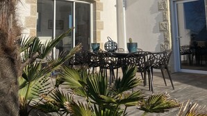 Un patio con zona de comedor, palmeras y un edificio con detalles de piedra y ventanas amplias.