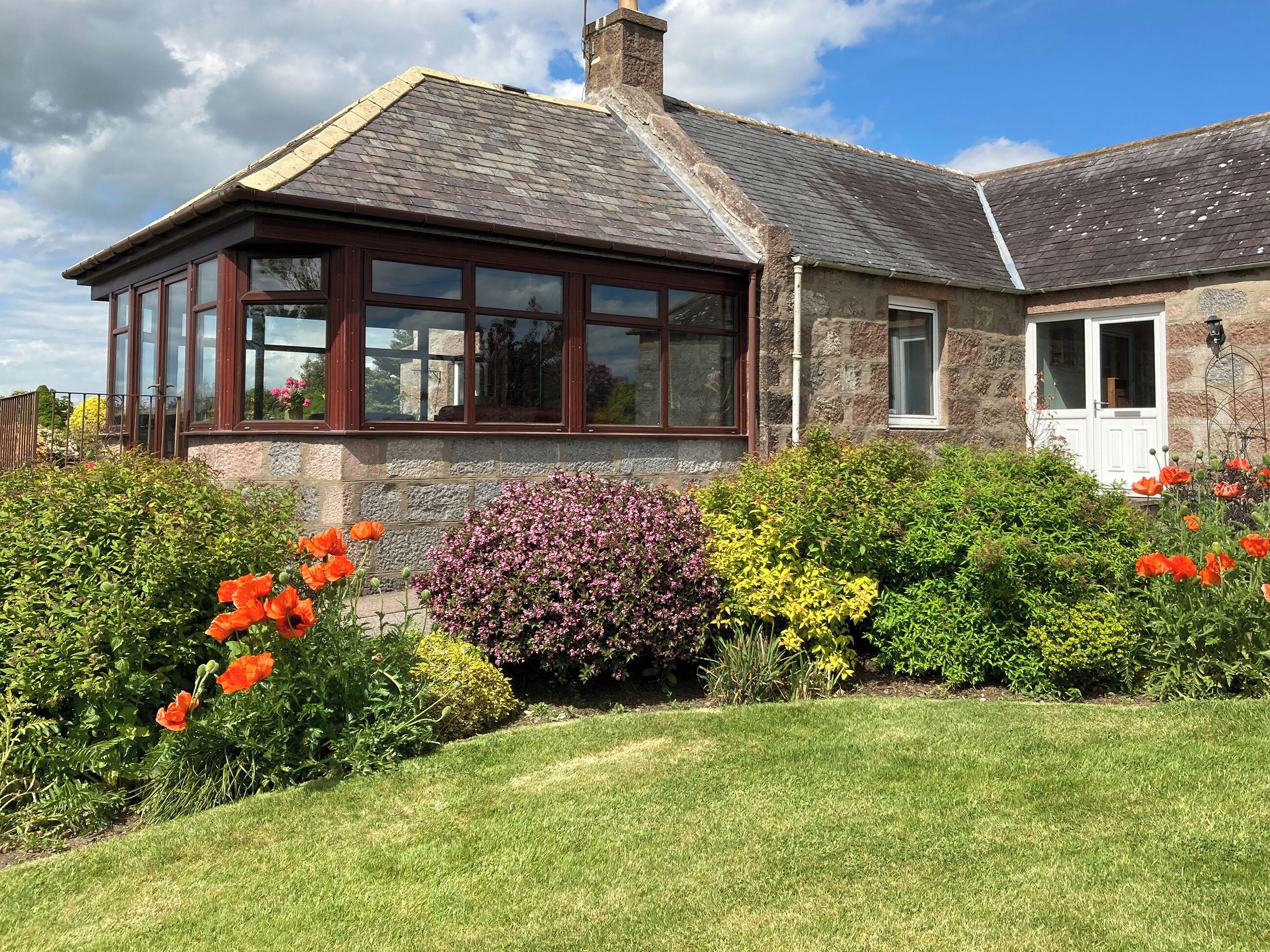 A house with a large bay window, stone walls, and a well-maintained garden with various shrubs and flowers.