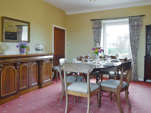 A dining room with a wooden table set for six, surrounded by six chairs. There is a wooden cabinet with a mirror and decorative items on top. A window with curtains is visible in the background.