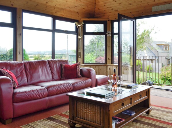 Una habitación con un sofá de cuero rojo, una mesa de centro de madera con libros y vasos, y ventanas amplias con vistas al exterior.
