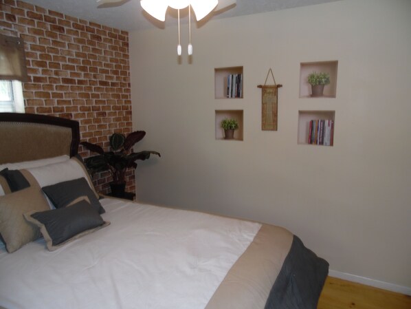 Un dormitorio con pared de ladrillo, cama con ropa de cama blanca y oscura, y estantes con libros y plantas.