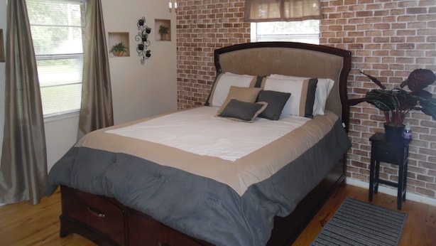 Une chambre avec un mur en briques, un lit avec une tête de lit, un ventilateur de plafond, une fenêtre avec des rideaux et une petite table sur laquelle est posée une plante.