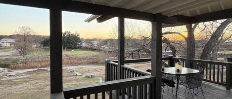 A covered outdoor deck with a table and chairs, overlooking a grassy area with trees and a distant view of a house.