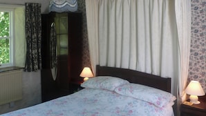 A bedroom with a bed, two bedside lamps, a window with curtains, a radiator, and a wooden wardrobe.
