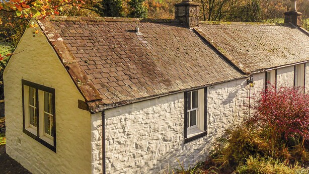 A stone house with a sloped roof and a chimney, surrounded by lush vegetation.