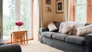 Una sala de estar acogedora con un sofá gris, una mesita de madera con un jarrón de flores y vistas a la vegetación a través de amplios ventanales.