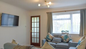 Uma sala de estar com televisão de tela plana na parede, um conjunto de sofás, uma porta com painéis de vidro fosco e uma janela com cortinas.