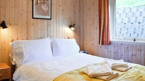 Un lit bien fait, avec des draps blancs et une couverture jaune, une table de chevet avec une lampe, et une fenêtre donnant sur un mur de briques.