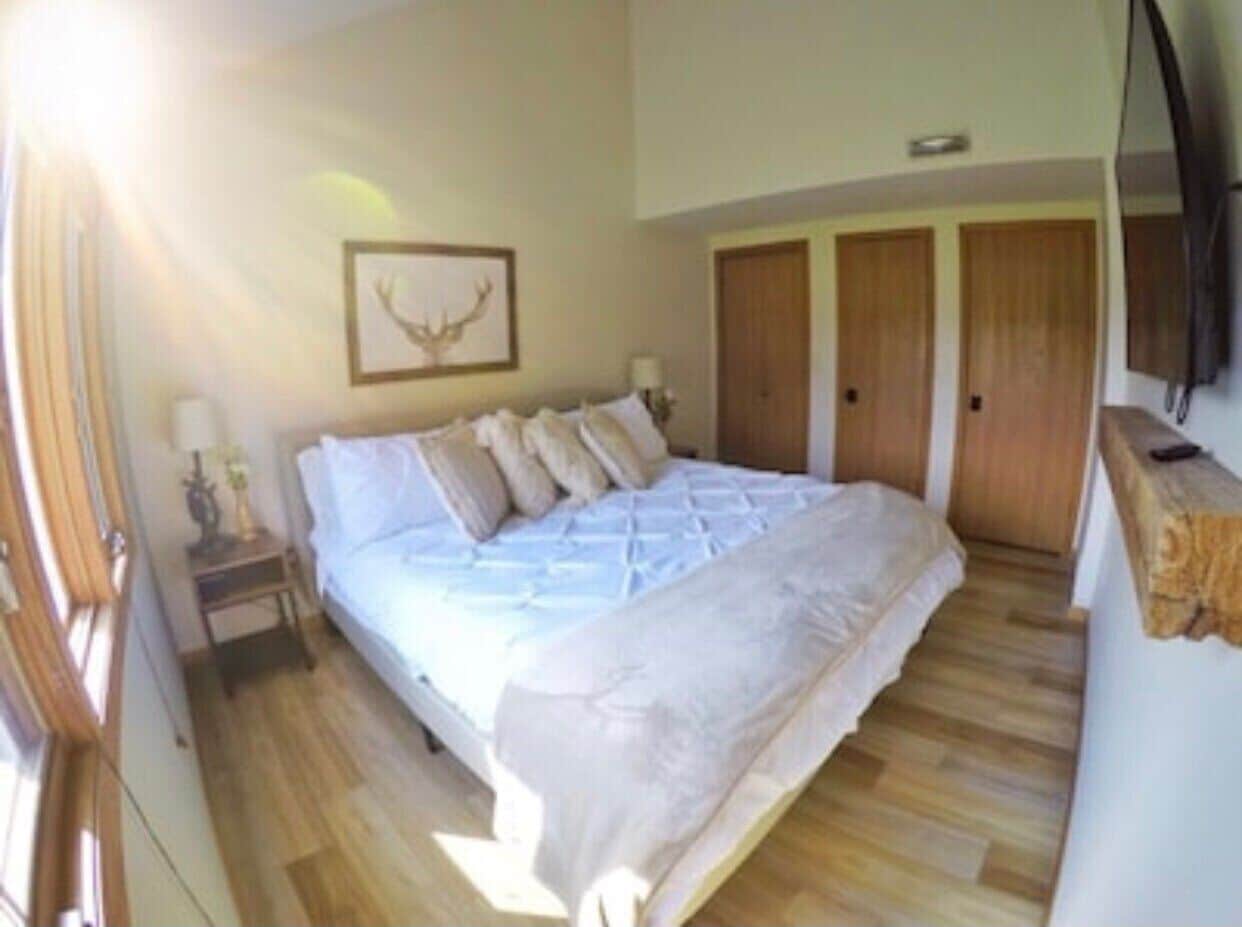 A bedroom with a large bed, wooden floors, a bedside table with a lamp, and a framed picture on the wall.