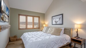 A bedroom with a large bed, a window with blinds, a framed deer head picture, a lamp on a nightstand, and a woven basket on the floor. - REMODELED! 2B/2B, Deck off10th GOLF Green, Dog Friendly, Pool, WIFI, in Resort! (Galena)