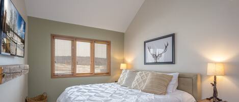 A bedroom with a large bed, a window with blinds, a framed deer head picture, a lamp on a nightstand, and a woven basket on the floor.