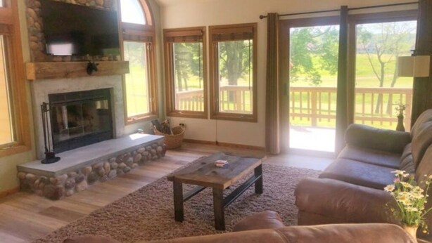 A cozy living room with a stone fireplace, comfortable seating, and large windows offering a view of the outdoors. - REMODELED! 2B/2B, Deck off10th GOLF Green, Dog Friendly, Pool, WIFI, in Resort! (Galena)