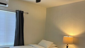 Quarto de hotel com duas camas, um ventilador de teto, ar condicionado, mesa de cabeceira com abajur e janela com persianas.