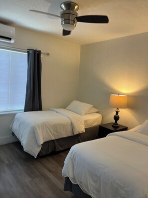 Une chambre d’hôtel avec deux lits, un ventilateur de plafond, un climatiseur, une table de chevet avec une lampe et une fenêtre avec des stores.