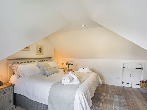 A bedroom with a sloped ceiling, a bed with white bedding, a bedside table with a lamp, a small bedside table with a vase of flowers, and a door with a basket on the floor.