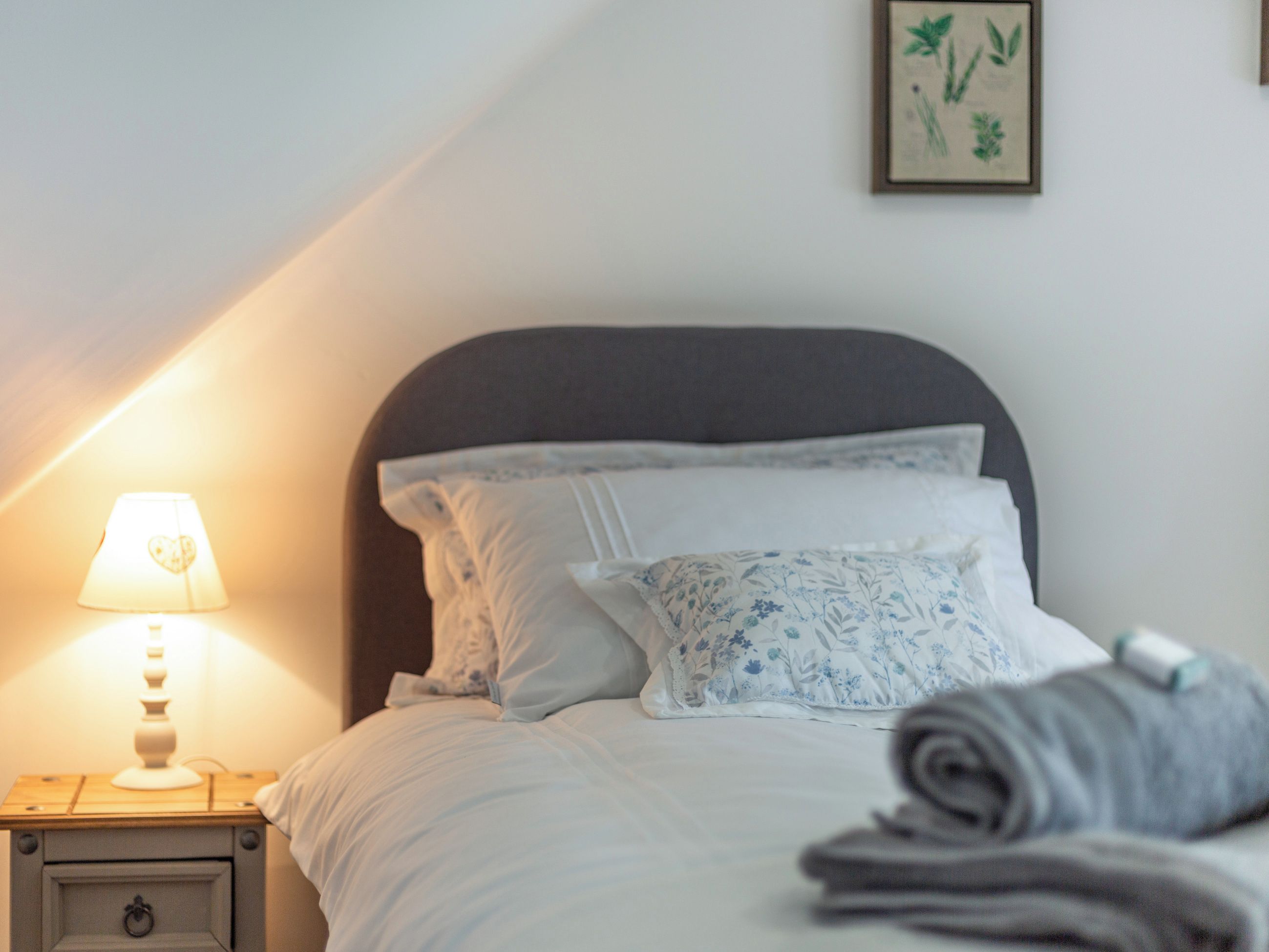 A neatly made bed with white and blue floral bedding, a bedside lamp, and a framed botanical print on the wall.