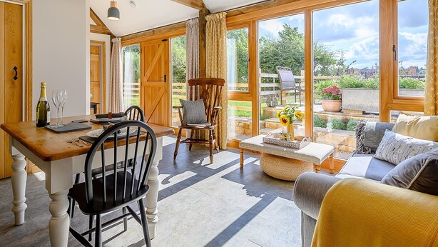 A room with a wooden dining table, chairs, a sofa, and a view of the garden through large windows.
