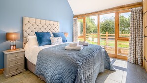 A bedroom with a large bed, a nightstand, a lamp, and a view of the outdoors through a large window.