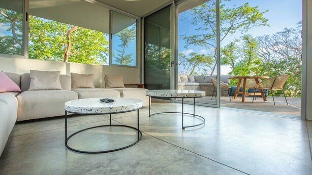 A modern living room with a sofa, two coffee tables, and a view of trees through large glass doors. - 2BR, 2.5BA, Jungle views, walk to beach, Pool, AC (Nicoya)